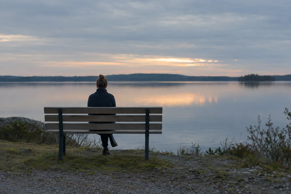 Person på en bänk vid en stilla sjö i skymningsljus, sedd bakifrån på avstånd